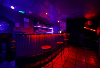 Interior of Cuero Cruising Club, a gay cruise club in Torremolinos. Bar with stools, red & blue lighting, and bottles on shelves.