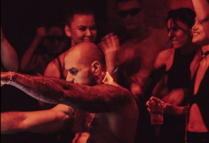 Gay Budapest nightlife: Shirtless man dancing at D9 Dark Nine club. Tattooed arm raised, other partygoers visible in the background. Red lighting.