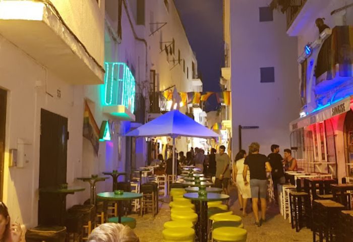 Ibiza gay bar, Dado Bar, at night. Outdoor seating on a narrow street. Rainbow flag visible. Gay travel advice, drinks, and nightlife.