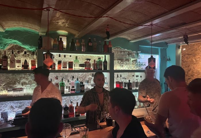 DADO Bar, a gay bar in Ibiza. Bartenders serve drinks to patrons. Shelves are stocked with liquor bottles. Nightlife scene.
