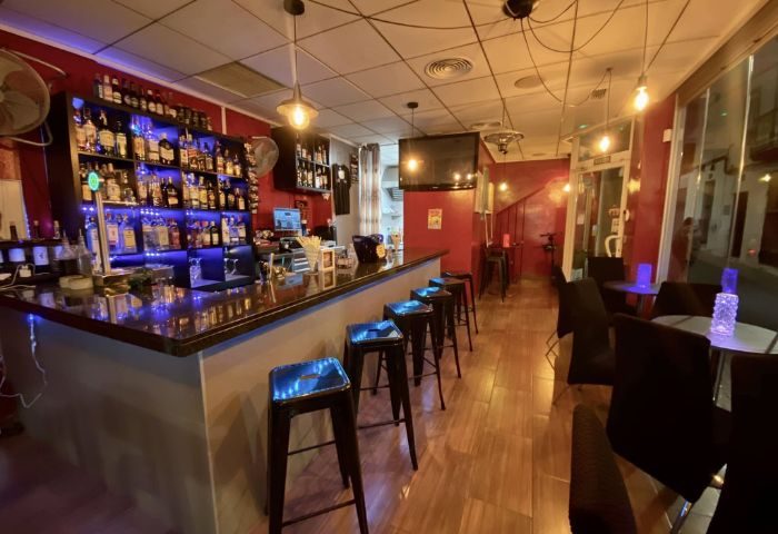 Interior of DD's Bar, a gay bar in Benidorm. Barstools line the counter, with tables & chairs beyond. Shelves stocked with liquor.