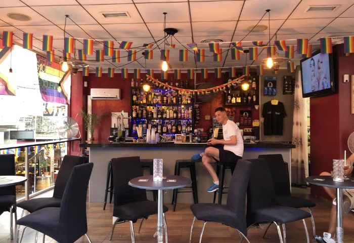 Interior of DD's Bar, a gay bar in Benidorm, Spain. Rainbow flags decorate the ceiling. A man sits at the bar with a drink.