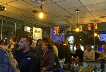 Interior of DD's Bar, a gay bar in Benidorm. People gather at the bar under warm lighting, with British and Spanish flags displayed.
