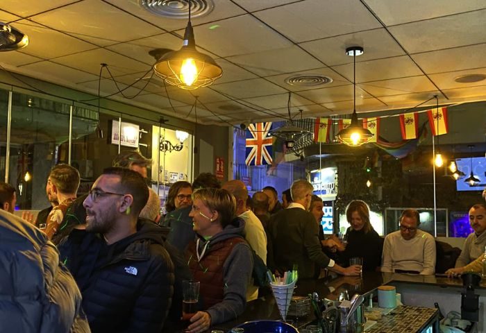 Interior of DD's Bar, a gay bar in Benidorm. People gather at the bar under warm lighting, with British and Spanish flags displayed.