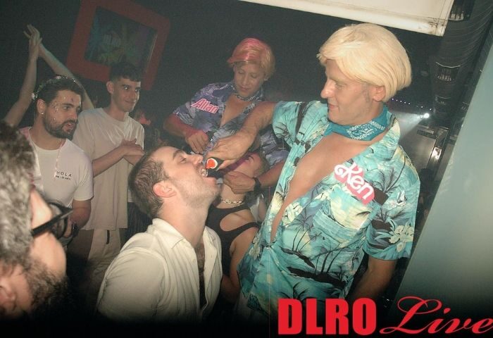 DLRO Live Gay Club in Madrid: Man pouring drink into another's mouth. Party scene with drag queens and crowd. Gay travel nightlife.
