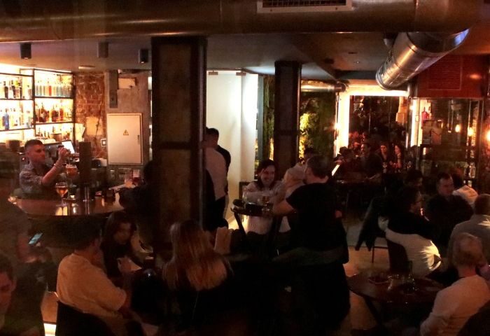 Interior view of a busy, dimly lit Mystic Gay Bar in Madrid. People are seated at tables and standing at the bar. Shelves of liquor are visible.