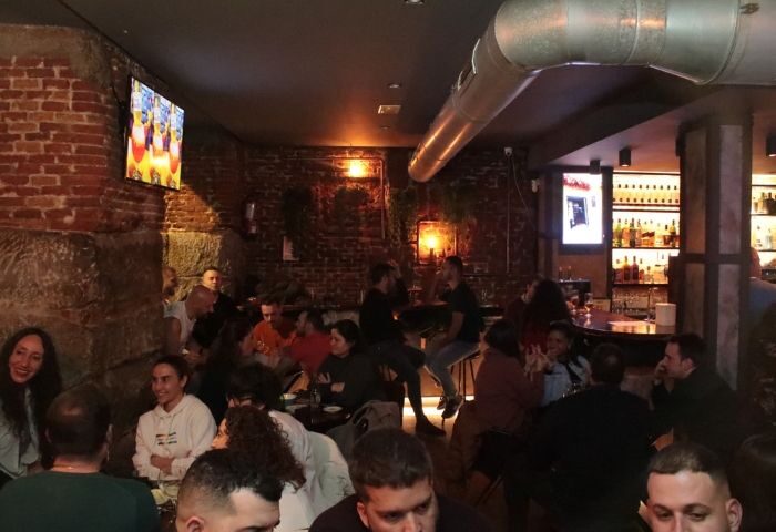 Cozy Mystic Gay Bar in Madrid. Patrons at tables and bar. Brick walls, warm lighting, and TV. Gay travel advice.