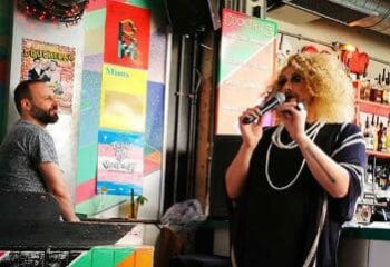 Dalston Superstore gay bar: Drag queen performing with a microphone. Man at the bar. Colorful wall art. Gay nightlife scene.