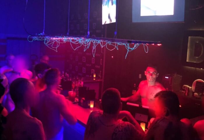 Depot Milano gay bar scene. Shirtless bartender serves patrons in a dimly lit, vibrant atmosphere. Gay travel advice, nightlife.