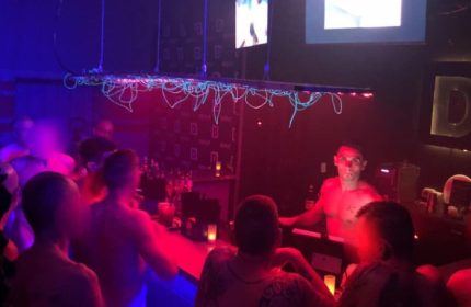 Depot Milano gay bar scene. Shirtless bartender serves patrons in a dimly lit, vibrant atmosphere. Gay travel advice, nightlife.