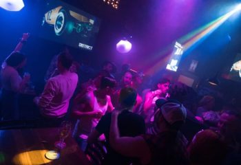 District Video Lounge, a gay bar in Montreal. Men socializing in a dimly lit space with video screens and colorful lights. Gay travel.