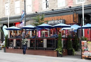 District Video Lounge, a gay bar in Montreal, features outdoor seating under blue umbrellas. People are enjoying drinks at the popular spot.