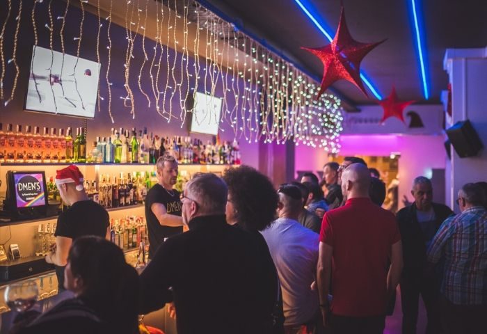Dmen's Gay Bar in Barcelona: Festive interior view with a crowd of gay men, barman, Christmas lights, and holiday decorations. Gay travel advice.