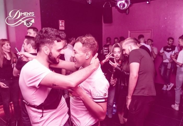 Dmen's Gay Bar in Barcelona: Two men embracing, smiling in a crowded, pink-lit gay bar. Nightlife scene, travel advice for gay men.