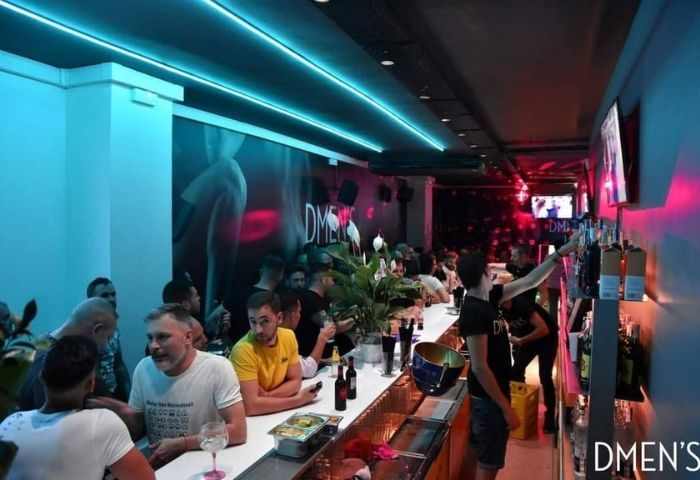 Dmen's Gay Bar in Barcelona. Interior view of the bar with people, blue lighting, and a bartender reaching for a bottle. Gay travel advice.