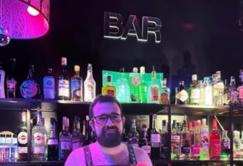 Drako Club Lisbon barman. Gay nightlife advice. Bartender with beard, glasses, leather harness stands before shelves of liquor bottles.