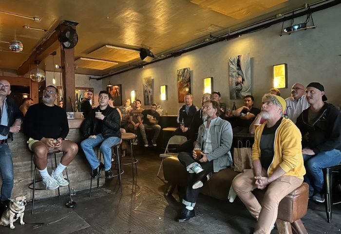 Gay men at Driftwood Bar in San Francisco watch a screen. The bar has a relaxed vibe, and they're enjoying drinks, a dog is present.
