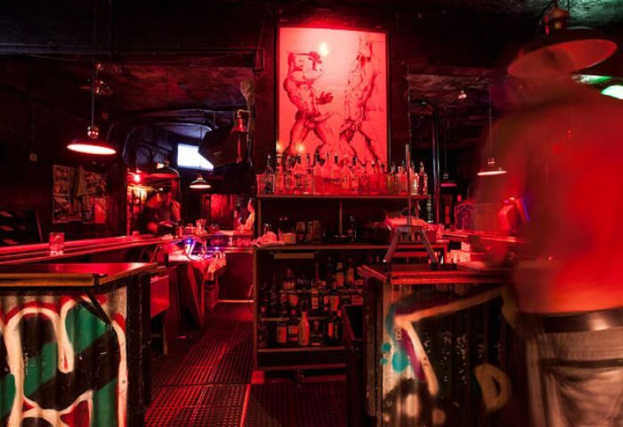 Interior of EAGLE LA gay bar. Red lighting illuminates the bar, bottles, and artwork featuring male figures. Graffiti decorates the bar front.
