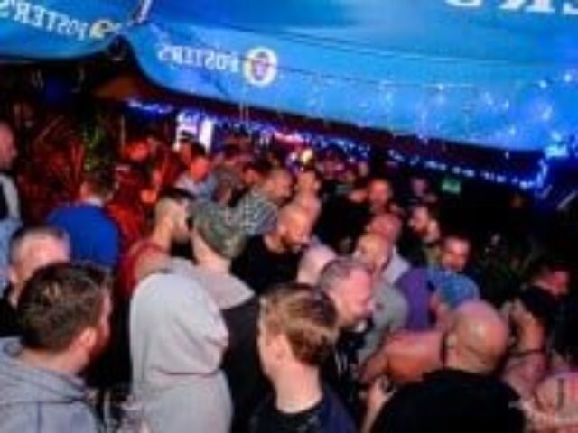 Inside EAGLE London, a lively gay club. A dense crowd of men, illuminated by blue lights, creates a vibrant, social atmosphere. Nightlife scene.