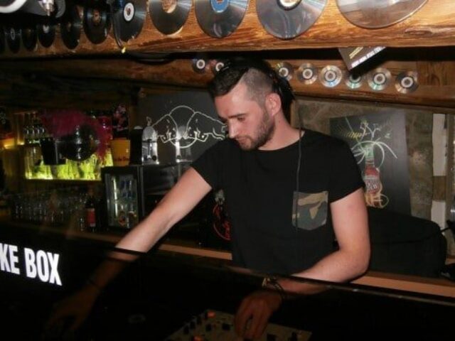 DJ at EAGLE London gay club. Man with undercut hairstyle, black t-shirt with camo pocket, at the soundboard. Vinyl records decorate the ceiling.