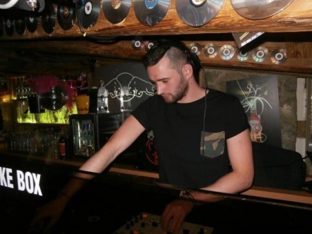 DJ at EAGLE London gay club. Man with undercut hairstyle, black t-shirt with camo pocket, at the soundboard. Vinyl records decorate the ceiling.
