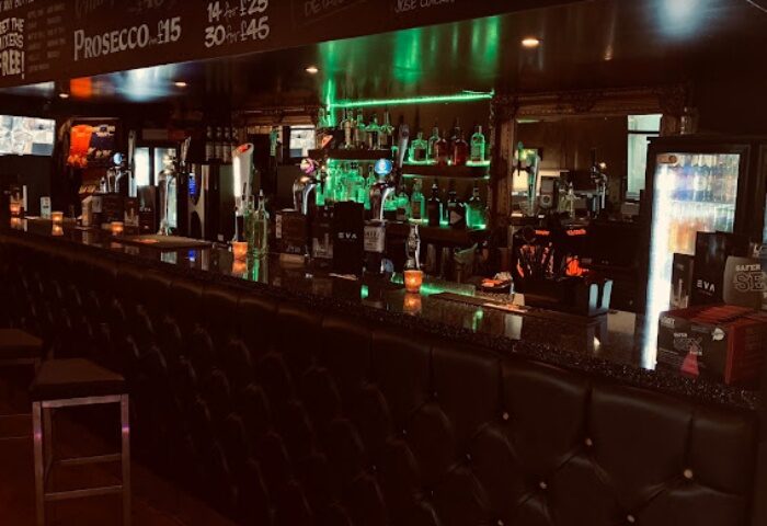 EVA Manchester bar interior. Bottles line the illuminated shelves behind the bar. A sleek, gay-friendly spot for drinks and socializing in Manchester's Village.