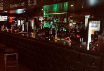 EVA Manchester bar interior. Bottles line the illuminated shelves behind the bar. A sleek, gay-friendly spot for drinks and socializing in Manchester's Village.