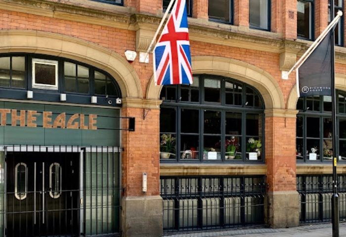 The Eagle Manchester, a gay bar, with a British flag. Brick building, arched windows, and dark trim. Gay travel advice.