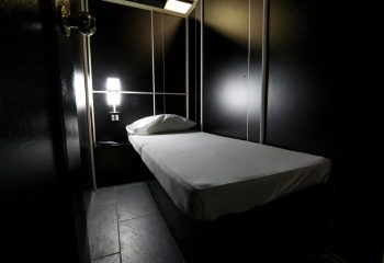 East Side Club, New York gay sauna. A private room with a bed, pillow, and nightstand. Dark walls and dim lighting create an intimate atmosphere.