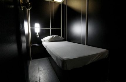 East Side Club, New York gay sauna. A private room with a bed, pillow, and nightstand. Dark walls and dim lighting create an intimate atmosphere.