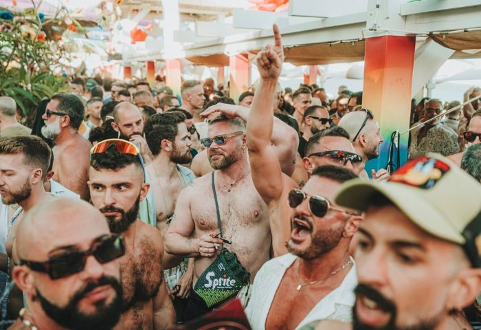 Gay men crowd at Eden Beach Club, a popular gay bar in Torremolinos, Spain. Party atmosphere, shirtless men, sunglasses, and rainbow decor.