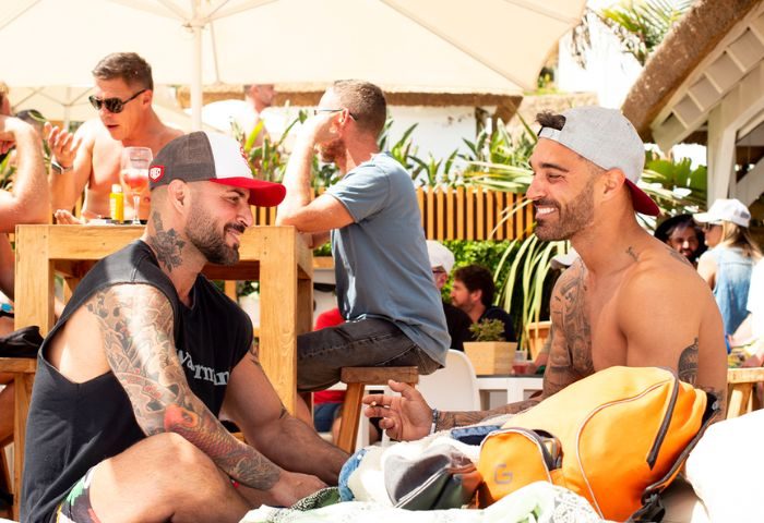 Gay men at Eden Beach Club, Torremolinos. Two tattooed men in caps smile at each other. Beach bar scene, gay travel, Spain.
