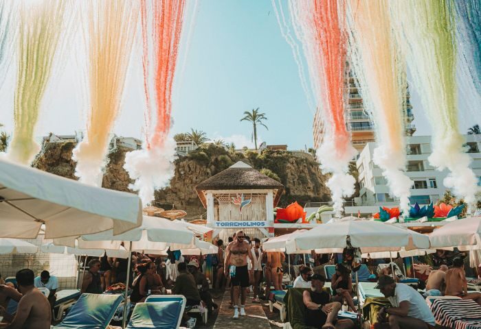 Eden Beach Club gay bar in Torremolinos. Beach scene with people under umbrellas, rainbow smoke effects, and the club's structure.