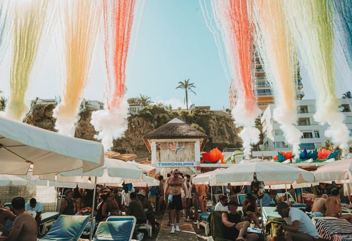 Eden Beach Club gay bar in Torremolinos. Beach scene with people under umbrellas, rainbow smoke effects, and the club's structure.