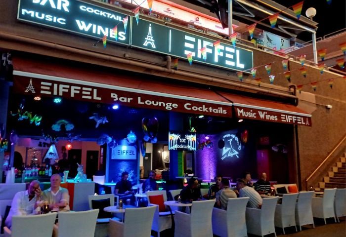 Eiffel Bar, a gay bar in Gran Canaria. Rainbow flags adorn the exterior. People sit at tables outside this popular gay travel destination.