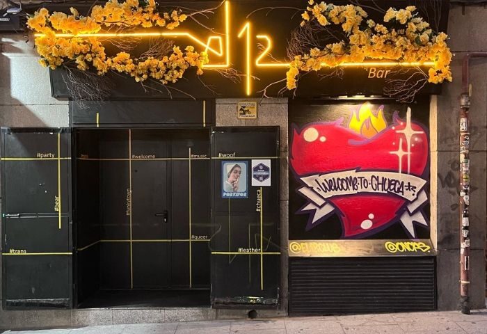 El 12 Club gay bar in Madrid. Entrance with neon sign, floral decor, and Welcome to Chueca heart graffiti. Gay nightlife travel.