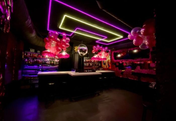 Madrid's El 12 Club gay bar interior. Neon lights, pink balloons, disco ball, and a well-stocked bar create a vibrant, welcoming atmosphere.