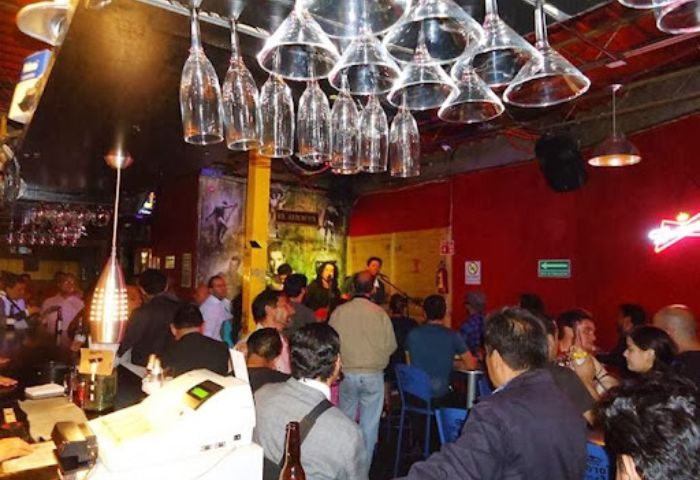 El Almacen Gay Bar in Mexico: Lively scene with patrons enjoying drinks and live music. Upside-down glasses hang above the bar.