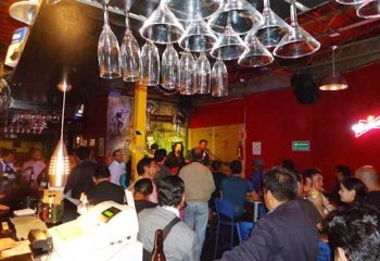El Almacen Gay Bar in Mexico: Lively scene with patrons enjoying drinks and live music. Upside-down glasses hang above the bar.