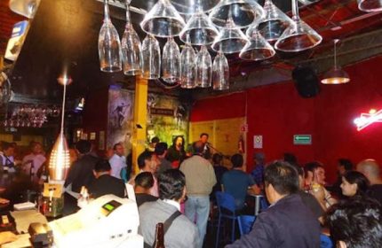 El Almacen Gay Bar in Mexico: Lively scene with patrons enjoying drinks and live music. Upside-down glasses hang above the bar.