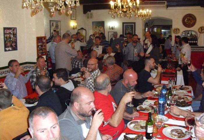 Interior view of El Horno Gay Bar in Sitges, Spain. Many gay men are seated at tables, eating and drinking. Chandeliers hang from the ceiling.