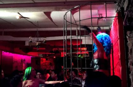 Nightlife at El Pecado Gay Bar in Mexico. Drag queen in cage, green-haired dancer, and crowd. Gay travel advice, gay bar scene.