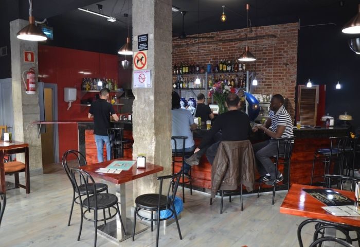 Interior of El Rincon Guay, a gay bar in Madrid. Patrons sit at the bar, with tables and chairs in the foreground. Brick wall detail.