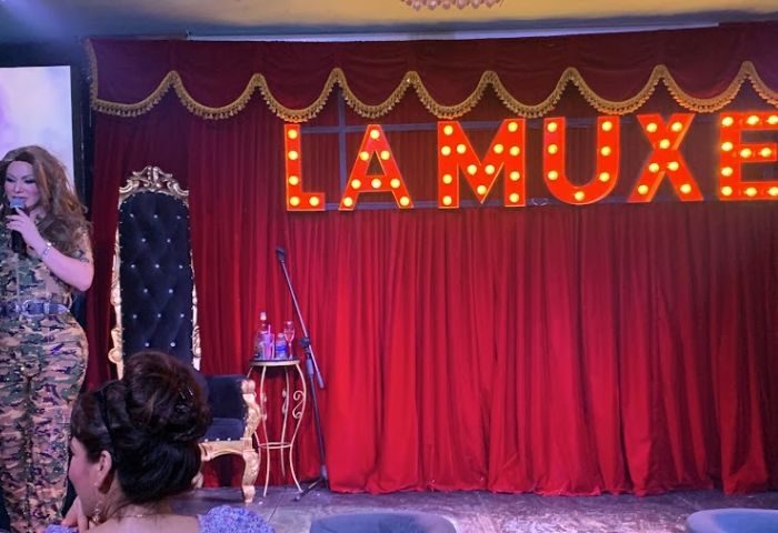 La Muxe gay bar stage in Puerto Vallarta. Drag queen with mic, red curtains, and illuminated sign. Gay travel destination.
