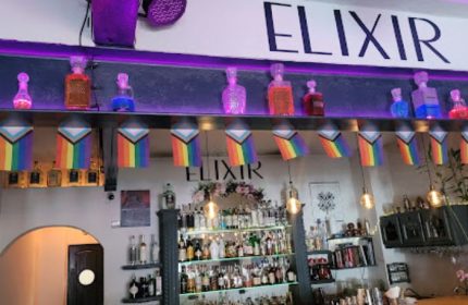 Elixir Mixology Bar interior in Puerto Vallarta. Gay party spot with rainbow flags, liquor bottles, and stylish decor. Nightlife scene.