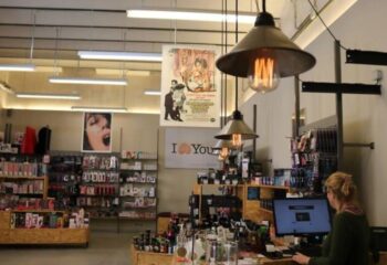 Inside Erotic Palace Barcelona, a gay cruise club. Shelves display adult items. Pendant lights hang above. Woman at computer.