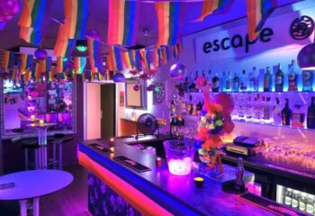 Escape Bar, a gay nightclub in Benidorm. Interior view with rainbow flags, bar with bottles, and neon lighting. Gay travel destination.