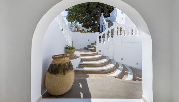 Carbonaki Hotel's archway view in Mykonos Town. Central location, white walls, stairs, and a large vase. Gay hotel travel destination.