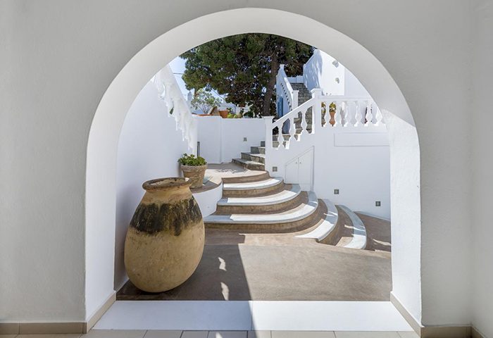 Carbonaki Hotel's archway view in Mykonos Town. Central location, white walls, stairs, and a large vase. Gay hotel travel destination.