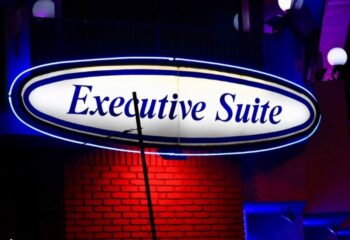 Executive Suite location: Neon sign for gay travel lodging. Upscale suite for gay men's vacation. Red brick wall, blue lighting.